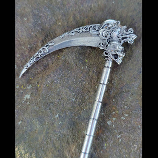 A full view of the Santa Muerte-inspired silver hair scythe, emphasizing the rich detailing of the floral engravings on the curved blade and the prominent skull at the top. The handle's segmented design draws the eye downward, with the craftsmanship capturing the essence of a ceremonial weapon, set on a textured stone surface.