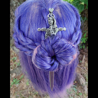 : A silver hair sword is secured in a braided purple hairstyle, its handle designed as an intricate depiction of the Norse goddess Hel. The top features a heart-shaped detail, and Hel is depicted with one half of her face skeletal and the other half human, symbolizing duality. The braid elegantly holds the sword in place, set against a natural outdoor background.