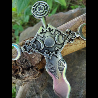 A side angle of the Hekate-inspired hair dagger, resting on a weathered branch, with visible engravings of torches, a triple moon sigil surrounded by a frame of four skeleton keys, and geometric shapes, exuding a vintage metallic finish.