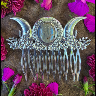 Front view of a silver hair comb placed amidst magenta flowers on a textured rock. The comb's features a triple moon and labradorite design stand out as the centerpiece, complemented by lush floral engravings and a mystical aura