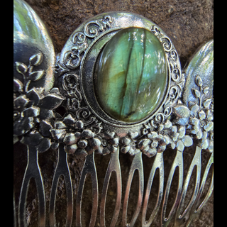 Focused view of the labradorite centerpiece of a silver hair comb, framed by scrolling silver patterns. The stone's iridescent green and blue tones are highlighted, reflecting light and enhancing its magical appearance