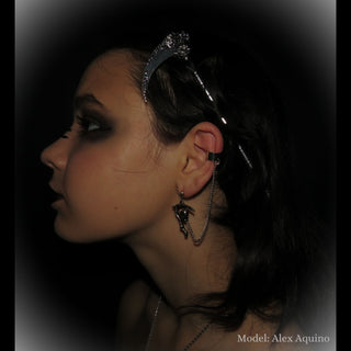 A side profile shot of a model wearing the Santa Muerte-inspired silver hair scythe as a hair accessory. The ornate floral blade curves above the head, with the skull detail adding a bold statement. The segmented handle adds texture, complementing the model's dark, dramatic styling and accompanying accessories, creating a cohesive gothic aesthetic.