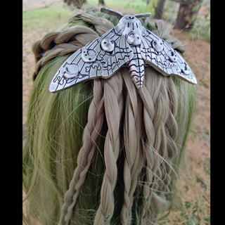 A silver moth-shaped hair clip with intricate lunar designs is pinned onto green and blonde braided hair. The moth's wings feature detailed crescent moons, lines resembling veins, and small circular accents, creating a celestial, mystical appearance. The lush outdoor setting adds a natural, earthy background to the piece.