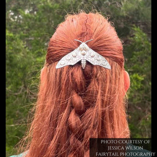 A vibrant red-haired model wears the silver moth hair clip against her braided hairstyle. The moth’s wings, adorned with celestial details, spread wide over her hair. The soft greenery in the background complements the mystical and elegant design of the hairpiece. Credit: Jessica Wilson of Fairytail Photography.