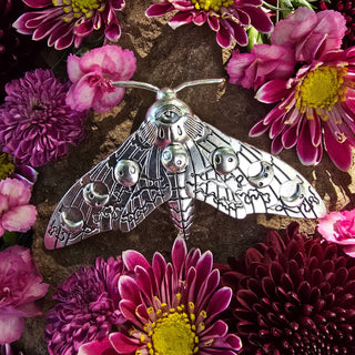  The silver moth hair clip is surrounded by a vibrant arrangement of purple and pink chrysanthemums. The intricate wing details, featuring crescent moons and celestial patterns, are accentuated by the floral arrangement, enhancing the magical, nature-inspired theme of the piece.