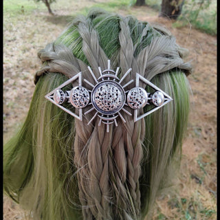 A silver-tone hair comb inspired by the phases of the moon is nestled in braided green hair. The intricate design includes a textured central sphere, flanked by crescents and geometric frames radiating outward. Pine needles scatter the background.