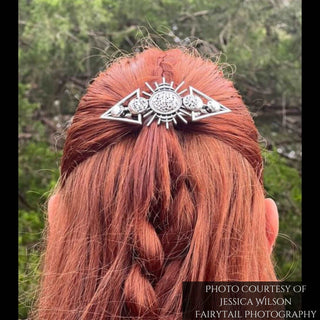 A model with vibrant red hair wears the celestial hair comb, secured above a neatly braided section. The silver finish of the comb contrasts against the natural background of lush green foliage