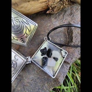 Close-up of an open diamond shaped locket pendant resting on a rock, containing small pieces of black and white stones inside. The locket has a black cord attached and features intricate celestial designs on the outside.