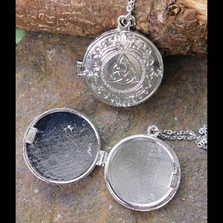 Close-up of a round silver locket with intricate Celtic designs, featuring a raised triquetra symbol surrounded by ornate scrolling patterns. The open locket displays two inner compartments, one textured in matte black and the other smooth silver, resting on a rugged stone backdrop with hints of bark texture.