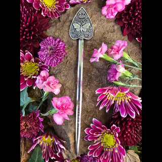 An intricately designed planchette-shaped hair stick, displayed vertically on a natural stone surface surrounded by vibrant pink and purple flowers. The metallic stick features a textured surface with a central moth design, its wings outstretched beneath an engraved eye symbol, accompanied by the words "Goodbye," "Yes," and "No." The composition emphasizes the mystical and spiritual essence of the design, with the surrounding flowers adding a touch of ethereal beauty.