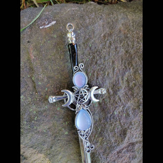 A silver hair stick adorned with a prominent triple moon sigil at the crest, with a central pentacle flanked by two crescent moons. Two opalescent stones, one oval and one teardrop-shaped, enhance the intricate design. The hair stick is positioned on a natural stone background, with grass faintly visible in the frame.