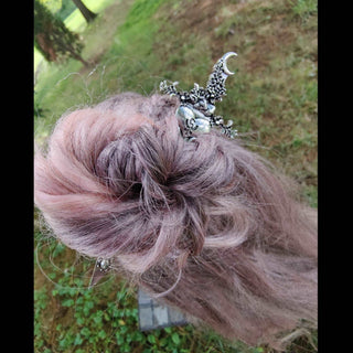 A hair dagger inspired by the Goddess Persephone, showcasing a crescent moon and intricate floral motifs extending from a silver heart design at the top. The dagger is securely nestled into an elegant, voluminous bun of soft, rose-tinted hair, with a natural outdoor backdrop of grass and trees adding to its mystical charm.