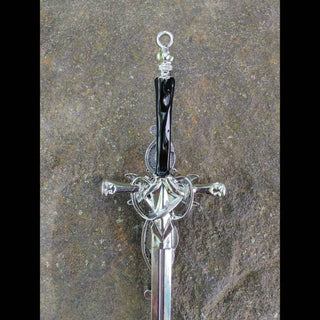 A silver hair stick featuring a detailed triple moon design at the crest, with a pentacle at the center. The handle is a polished black resin, wrapped with coiled silver wire at the top, ending in a loop for decorative use. The hair stick is displayed on a rough-textured stone surface, emphasizing its reflective silver finish.