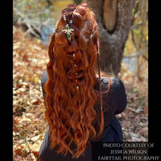 Rear view of long, flowing red hair styled with a complex braid pattern, showcasing a green gemstone sword-shaped hair stick placed against an autumnal forest backdrop with fallen leaves and golden sunlight