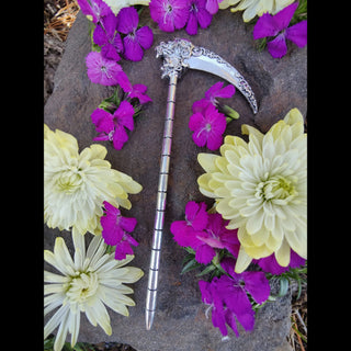 The Santa Muerte-inspired silver hair scythe displayed on a bed of vibrant purple and pale yellow flowers. The curved blade, with its floral engravings, and the skull detail at the top stand out amidst the soft floral setting. The segmented handle draws the eye, blending the scythe's haunting beauty with the delicate natural background.