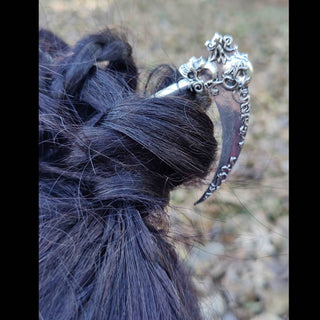 A Santa Muerte-inspired silver hair scythe styled in dark hair, emphasizing its intricate details. The skull motif at the top and the floral engravings along the blade create a striking contrast against the hair, evoking a sense of gothic mystique. The handle's segmented design enhances the scythe's realism in its functional yet decorative role.