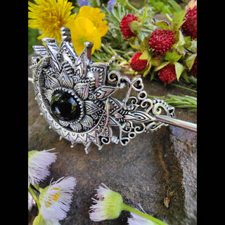 Side angle of a crescent moon hair stick showcasing intricate Victorian-inspired patterns with a black gemstone centerpiece, placed on a rugged stone background amidst dandelions, buttercups, wild strawberries, and soft white blossoms