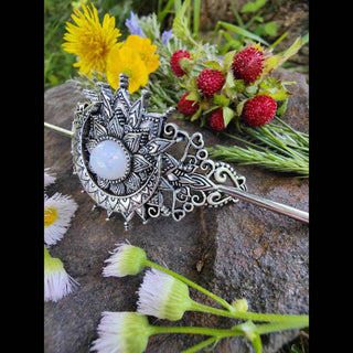 Close-up of a crescent moon hair stick with ornate Victorian details and an opalescent stone centerpiece, displayed among vibrant wildflowers and textured greenery on a rugged stone background