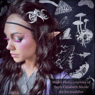 A model styled with elf ears and fantasy-inspired makeup, adorned with the raven skull and crescent moon barrette. The background features whimsical mushroom and forest illustrations, perfectly complementing the mystical theme.