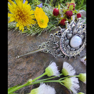 Side view of a crescent moon hair stick showcasing its detailed Victorian-inspired metalwork with a glowing opalescent stone centerpiece, placed on a textured stone surface surrounded by bright yellow dandelions, delicate lavender sprigs, and red wild strawberries