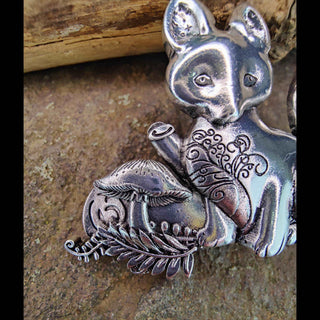 Detailed close-up of a silver fox hair barrette, highlighting the engraved mushroom and leaf designs beside the fox, with fine etchings and natural textures in the background