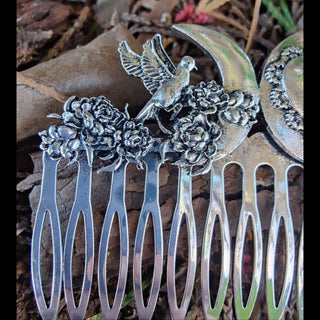 A side angle of the hair comb showing the crescent moon and bird details with clustered flowers, set against a backdrop of earthy tones and leaves, emphasizing its organic inspiration