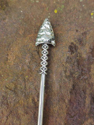 Close-up of the engraved arrowhead of a silver hair stick with intricate detailing, displayed on a natural stone background.