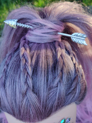 Silver arrow-shaped hair stick styled in a high braided bun on lavender-colored hair, shown in an outdoor setting