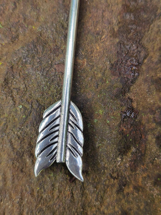 Close-up of the feathered end of a silver arrow hair stick, displayed on a textured stone surface.