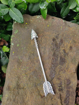 Full view of a silver arrow hair stick with detailed arrowhead and feather engravings, displayed on a textured stone surface surrounded by greenery.