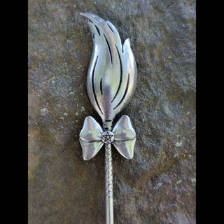 Close-up of a silver hair stick designed like a broom with textured bristles and a bow detail, displayed on a natural stone background