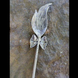 Full view of a silver broom-shaped hair stick featuring a textured bristle design, a bow with a pentagram engraving, and engraved moon phases on the handle, displayed on a stone background