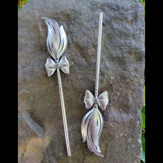 Pair of silver broom-shaped hair sticks, one shown with bristles pointing upward and the other downward, displayed on a textured stone surface