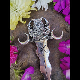 Full view of a silver hair stick featuring an owl perched between crescent moons, with a flowing, wavy design and engraved arrows, styled on a natural stone surface surrounded by colorful flowers