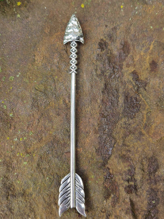 Full view of a silver arrow hair stick with detailed arrowhead and feather engravings, displayed on a textured stone