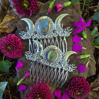 Two silver hair combs featuring a triple moon sigil centered with labradorite stone arranged together on a rock. The surrounding magenta and purple flowers create a vibrant, enchanting setting, highlighting the intricate craftsmanship and magical theme of the combs. 