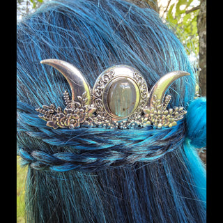 Close-up of a silver hair comb with intricate floral details and a central labradorite stone. The comb features a crescent moon motif on both sides of the stone, surrounded by ornate designs and delicate flowers. It is woven into braided, vibrant blue hair with subtle teal highlights, creating a celestial and enchanting look.