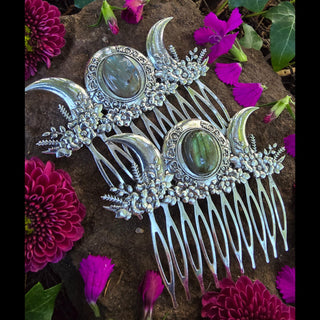 Two identical silver hair combs displayed on a rock surface surrounded by magenta flowers. Each comb showcases crescent a triple moon design. the moons on either side of a polished labradorite stone, with intricate floral decorations extending along the base, exuding a whimsical and ethereal vibe.