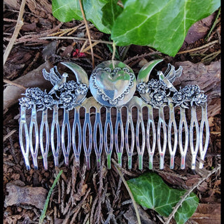 The entire silver hair comb resting on forest floor foliage, showcasing its intricate design with crescents, a central heart, and floral motifs. The bird embellishments add a sense of movement and grace.