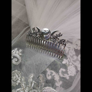 A silver decorative hair comb placed on a sheer white lace veil. The comb features a crescent moon design on both ends, a central heart motif, and floral detailing, exuding a romantic and celestial vibe