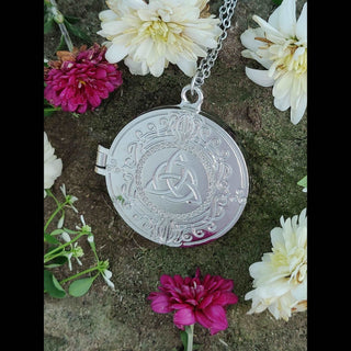 Silver Celtic locket laid on a mossy rock surrounded by small white and magenta flowers. The locket's triquetra design and ornate border details gleam against the natural greenery, with the vibrant petals adding pops of color to the composition.