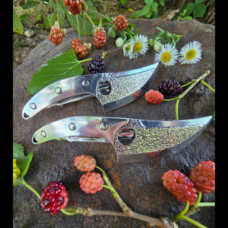 Set on a natural stone surface with ripe blackberries and daisies, the two knife hair clips create a rustic yet sharp visual statement