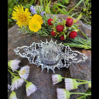 The back of a crescent moon-shaped hair stick featuring a Victorian-inspired design with intricate filigree and a sturdy clasp, displayed on a rugged stone background surrounded by freshly picked dandelions, buttercups, and wild mock strawberries