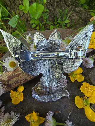 back view of a silver wing-shaped hair clip, highlighting the intricate etched texture of the wings, displayed on a textured stone surface with scattered buttercup blossoms and delicate white wildflowers, set against a natural forest backdrop