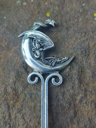 A close-up of a silver dragon perched on a crescent moon, with intricate scales and wings visible, against a rocky background.