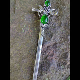 Close-up of a sword-shaped hair stick with intricate filigree, a central green teardrop gemstone, crescent moon details, and a pentacle design, displayed on a textured stone surface with subtle moss accents