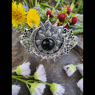 Full view of a crescent moon-shaped hair stick featuring a black gemstone centerpiece and intricate filigree design, displayed on a stone surface surrounded by wild dandelions, buttercups, wild strawberries, and white blossoms