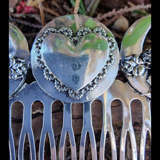 A close-up of the silver hair comb's central heart motif. The heart is encircled with delicate floral engravings, reflecting a vintage and elegant aesthetic