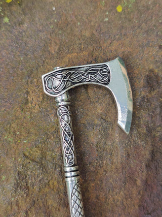 Detailed close-up of a silver Viking hatchet hair stick featuring Celtic knot engravings, photographed on a stone surface