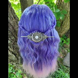 Crescent moon hair stick securing styled lilac-purple hair with loose curls, worn in an outdoor setting surrounded by lush greenery and tree trunks, highlighting the luminous opalescent stone and intricate Victorian-inspired design